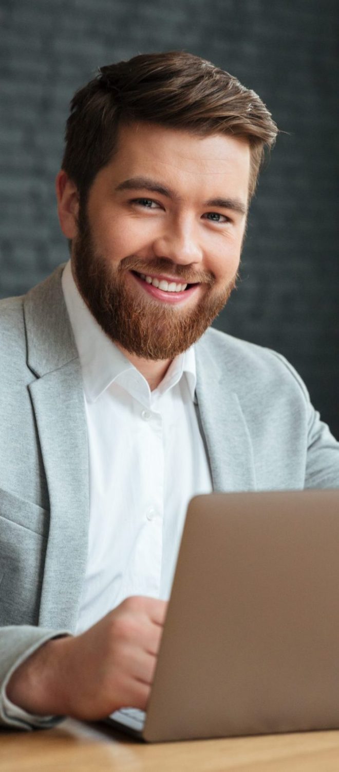 Image of cheerful young caucasian businessman sitting indoors using laptop computer. Looking camera.